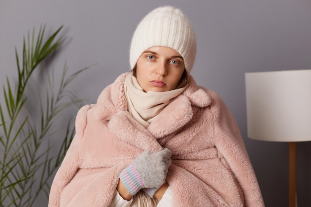 Indoor shot of pretty sad upset woman wearing cap and coat posing in living room being in depression feels cold can not to get warm in cold apartment