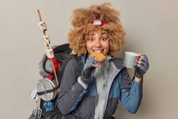 Indoor shot of pretty woman hiker has snack during journey\
bites waffle and drinks tea dressed in comfortable clothes carries\
backpack isolated over grey background female adventurer enjoys\
picnic