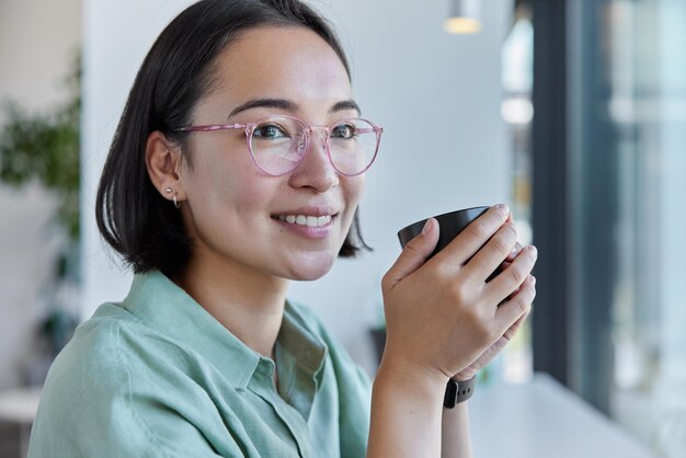 Indoor shot of pretty young asian woman with dark hair has\
cheerful satisfied dreamy expression holds mug of tea or coffee\
looks away happily wears spectacles and shirt being deep in\
thoughts