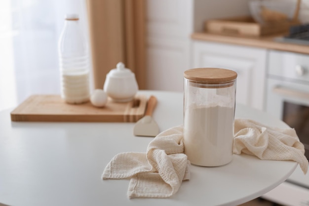 Indoor shot of products for baking, milk in the bottle, sugar, egg, cutting board, sugar, kitchen spatula, flour in a jar and dish towel standing on the table with white kitchen set on background.