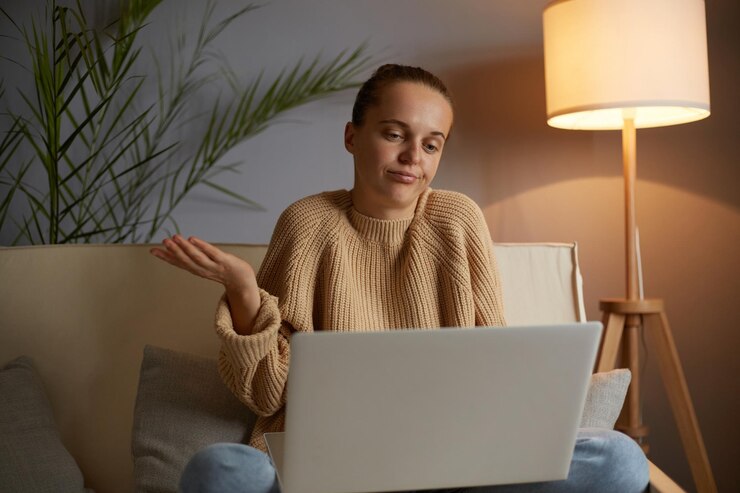 Photo indoor shot of puzzled confused caucasian woman wearing beige jumper and jeans sitting on cough in lotus pose having video call on laptop shrugging shoulders looks helpless