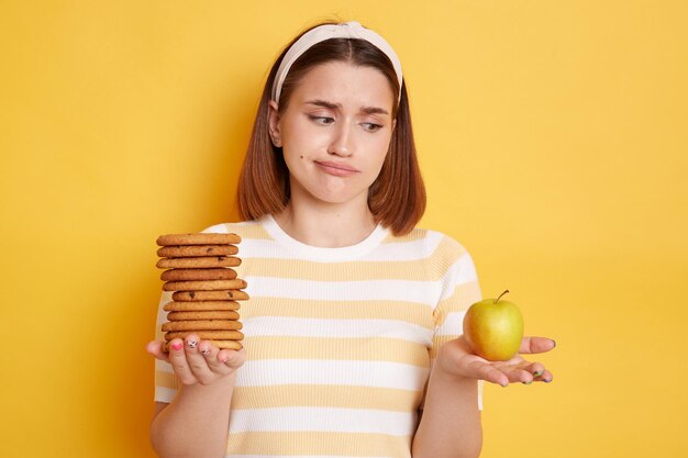 Indoor shot of puzzled confused woman wearing t shirt and hair band posing isolated on yellow background holding cookies and apple doesn't know what to choose healthy or junk food