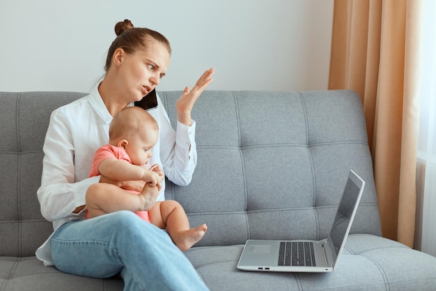 Indoor shot of puzzled confused woman with hair bun wearing white shirt and jeans, working online during maternity leave, talking on phone with partner, sitting on sofa with infant baby daughter.