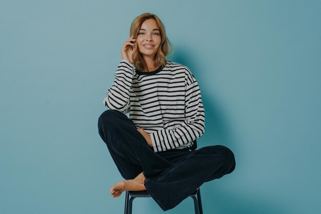 Indoor shot of relaxed woman with gentle smile on face sits crossed legs bare feet on chair