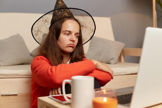 Indoor shot of sad bored caucasian woman in witch hat having\
online video call on laptop celebrating halloween alone at home\
looking at display with pout lips being in bad mood