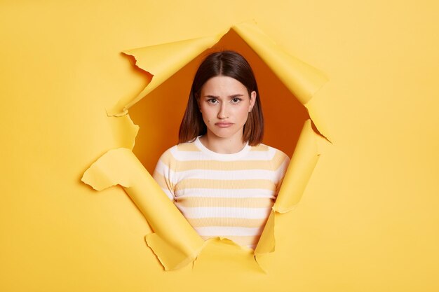 Indoor shot of sad upset attractive woman stands in torn paper hole looking at camera with pout lips and expressing sorrow looking through breakthrough of yellow background