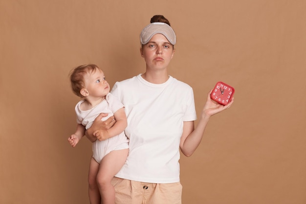 Indoor shot of sad upset Caucasian woman with dark hair wearing sleeping standing with her baby daughter in hands and showing alarm clock sleepy mother needs more rest