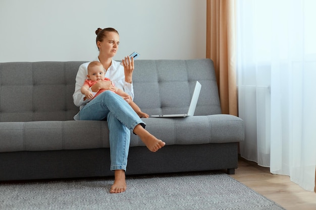 Premium Photo | Indoor shot of sad upset woman with hair bun wearing white  shirt and jeans sitting on sofa with infant baby daughter, working online  during maternity leave, holding phone in