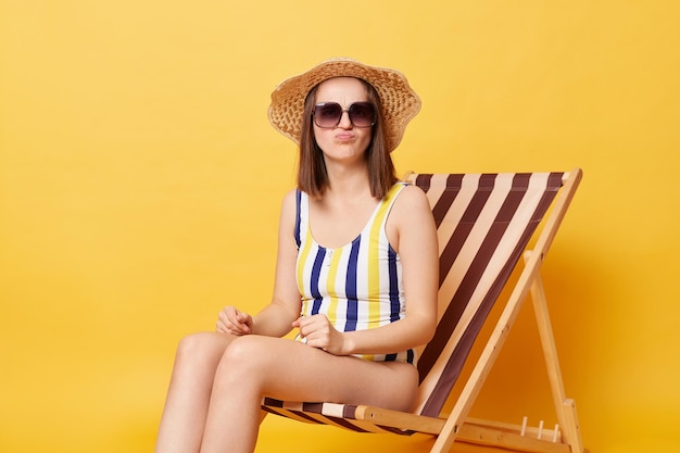 Indoor shot of sad upset woman with slim body wearing straw hat sunglasses and swimsuit posing isolated over yellow background looking at camera with pout lips being disappointed of her vacation