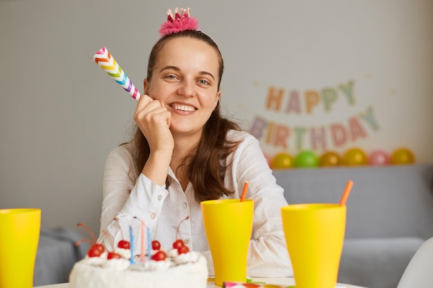 Indoor shot of satisfied delighted woman wearing white clothing sitting at table with cake and drink, looking at camera with happy charming smile, holding party horn.