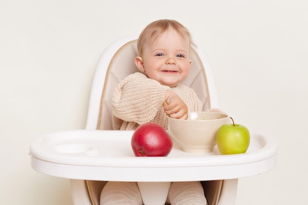 베이지색 스웨터를 입고 높은 의자에 앉아 흰색 배경에 격리된 접시에서 과일 퓌레를 먹는 만족하고 긍정적인 어린 소녀의 실내 사진