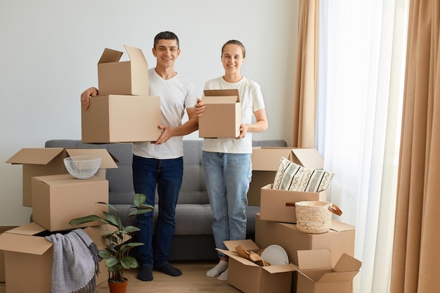 Внутренний снимок довольной позитивной молодой пары, сидящей на диване в окружении картонных коробок семьи во время переезда в новую квартиру, держащей личные стопки в картонных посылках