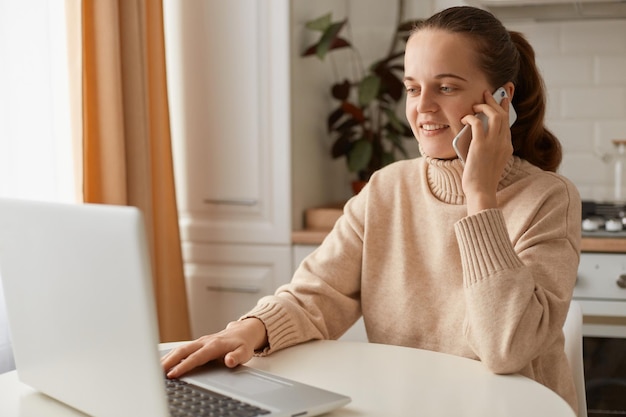 Indoor shot of satisfied woman wearing beige jumper posing in kitchen female working on notebook and talking via mobile phone having pleasant conversation smiling