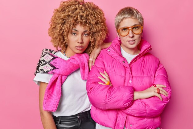 Indoor shot of self confident serious women look directly at\
camera dressed in stylish clothes spend time together being best\
friends listen information attentively isolated over pink\
background