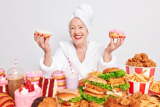 Indoor shot of senior woman smiles broadly and holds two delicious doughnuts has happy mood eats junk food