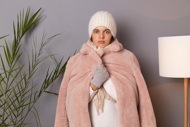 Indoor shot of serious attractive caucasian woman wearing cap\
and coat posing in living room without central heating feels cold\
wrapped in warm clothing