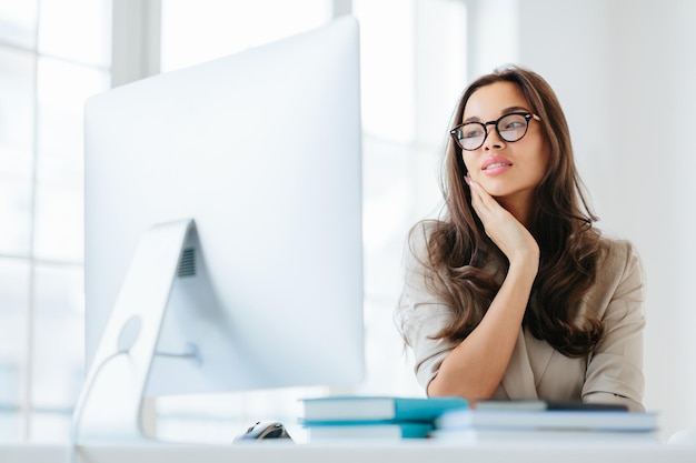 Indoor shot of serious brunette lady focused in monitor of computer uses wireless internet for publication keeps hand on cheek has attentive look sits at desktop works on business contract