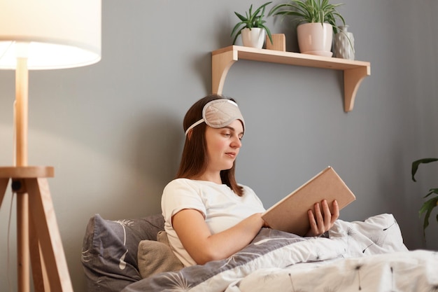 Indoor shot of serious concentrated brown haired woman in white tshirt and sleeping mask holding book in hands reading novel or poems being at home in bed