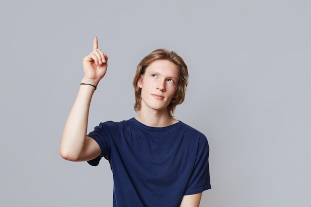 Indoor shot of serious man with trendy hairdo looks up and raises index finger