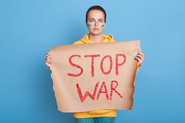 Indoor shot of serious patriotic woman in hoodie with Ukrainian flag on cheeks posing isolated over blue background protesting on demonstration with poster stop war