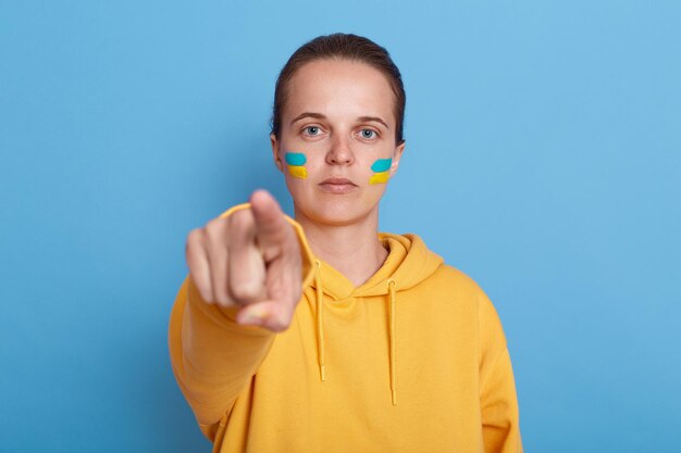 Indoor shot of serious patriotic woman wearing hoodie with blue and yellow flag on cheeks looking and pointing finger to camera Russian you can stop the war posing isolated over blue background