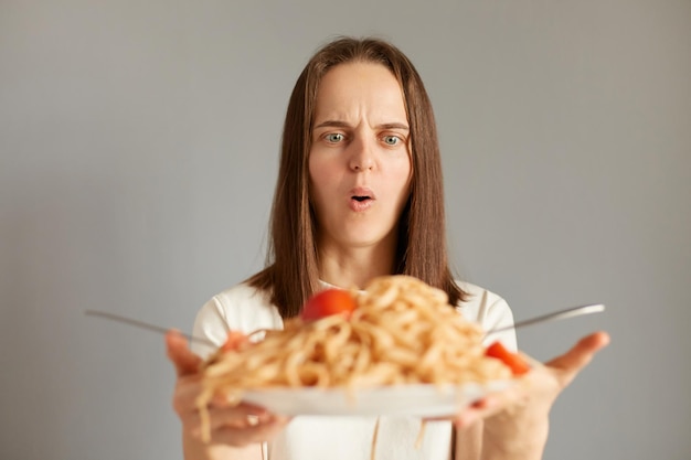 Foto colpo al coperto di una donna scioccata e stupita in maglietta bianca con in mano un piatto grande con tagliatelle che guardano con gli occhi grandi e la bocca aperta mantiene la dieta non può mangiare la pasta