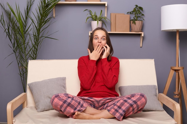 Indoor shot of shocked astonished brownhaired female wearing red sweater and checkered pants sitting on cough in room at home talking on smart phone hearing breaking news covering mouth with palm