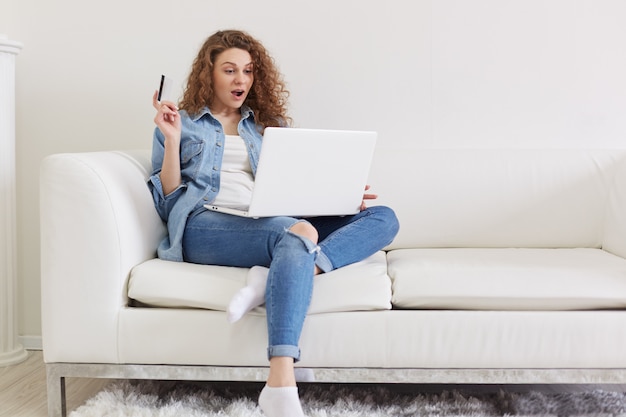 Indoor shot of shocked emotional lady with curly hair sitting in light living room