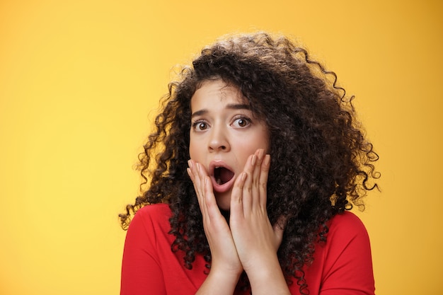 Indoor shot of shocked female drama queen with curly hair gasping in surprise, feeling empathy and fear turning head holding hands on cheeks astonished as hearing shook stunning news. Emotions concept