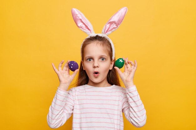 Indoor shot of shocked surprised girl with rabbit ears on her head with eggs isolated on yellow background looking at camera with big eyes sees something astonishing