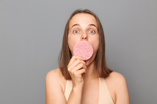 Indoor shot of shocked woman with brown hair and no makeup standing and covering mouth with sponge looking at camera with big eyes doing cosmetic procedures standing isolated over gray background