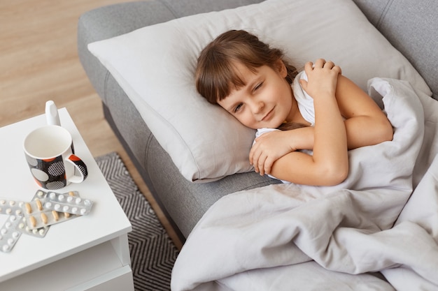 Ripresa al coperto di una bambina malata sdraiata sotto la coperta con le braccia incrociate e che guarda tristemente la telecamera mentre è sdraiata sul divano e soffre di alta temperatura e influenza o grippe.