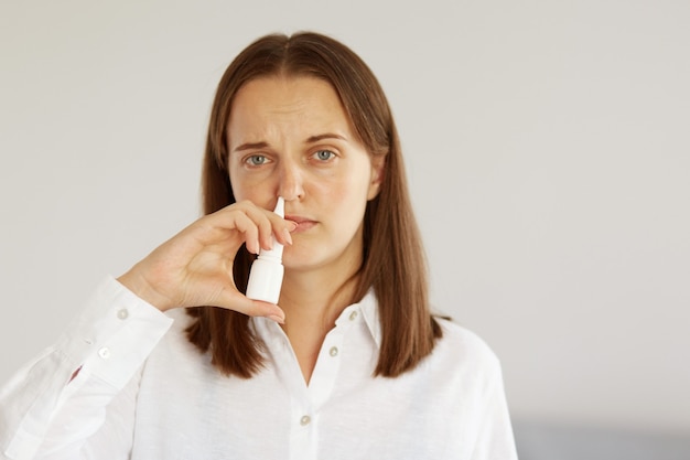 Indoor shot of sick winsome female with pleasant appearance wearing white casual style shirt, using nasal spray, suffering runny nose, looking at camera with ill frowning face.