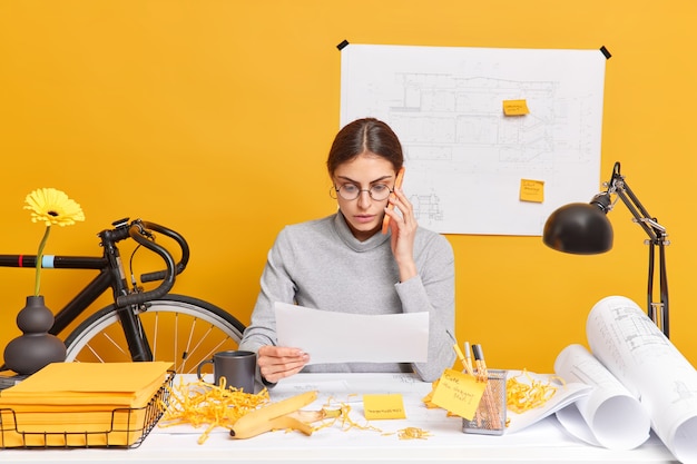 Indoor shot of skilled female architect discusses ideas for engineering project via smartphone concentrated in paper has mess at workplace involved in working process tries to finish project on time