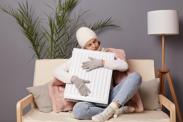 Indoor shot of sleepy frozen woman in coat and hat sit in cold\
living room and embracing radiator feels freeze at home having nap\
while getting warm keeps eyes closed