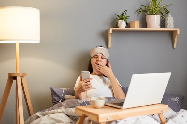 Indoor shot of sleepy tired woman wearing sleep mask sitting\
with laptop in bed at home holding mobile phone in hand looking at\
screen yawning and covering mouth with hand