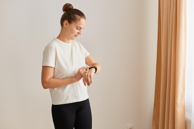 Indoor shot of slim attractive woman is using smart watch during workout at home, looking at her fitness tracker, checking indicators after workout, wearing white t shirt.