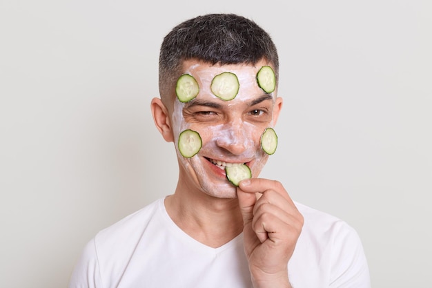 Indoor shot of sly cunning man wearing white t shirt taking\
care of his face doing procedures for nutrition and hydration of\
skin looking at camera and biting slice of cucumber