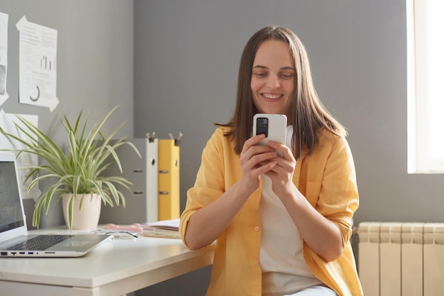 Indoor shot of smiling adorable woman with brown hair wearing casual outfit sitting in office and using cell phone expressing positive emotions having break at work