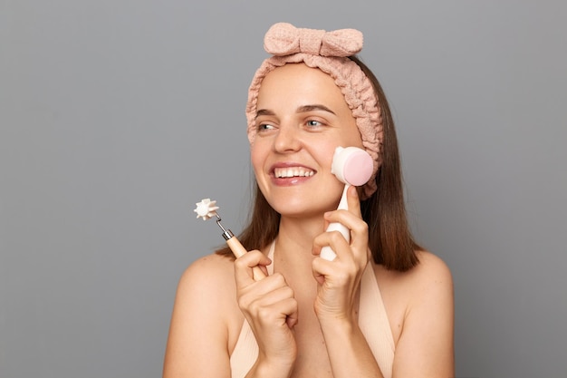 Indoor shot of smiling attractive beautiful woman with hair band holding gua Sha jade brush and massage roller looking away with charming smile isolated over gray background