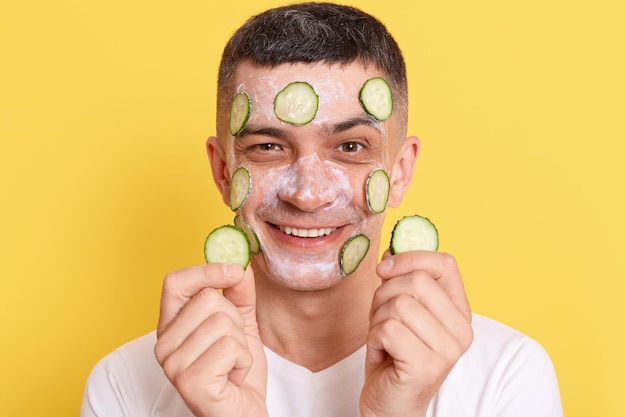 Indoor shot of smiling attractive man wearing white t shirt posing with mask and slices of cucumbers on his face expressing positive emotions enjoying spa procedures