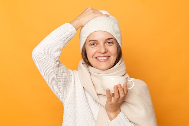 Indoor shot of smiling attractive young adult woman wearing white sweater hat and scarf posing isolated over yellow background looking at camera with satisfied face drinking hot coffee