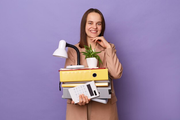 Indoor shot of smiling beautiful woman wearing beige jacket holding lot of documents folders looking at camera with toohy smile keeps hand under chin posing isolated over purple background