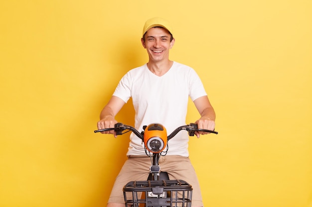 Indoor shot of smiling cheerful man wearing white t shirt and cap riding electric scooter having fun alone while having free time isolated on yellow background