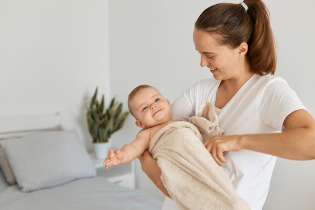 흰색 티셔츠를 입은 웃고 있는 검은 머리 여성의 실내 사진은 아기 딸을 손에 들고 수건으로 싸인 아이, 샤워를 한 직후 아이를 안고 서 있습니다.
