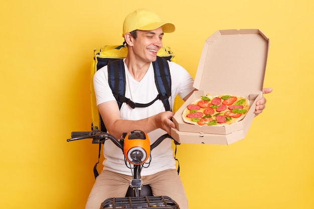 Scatto al coperto di sorridente felice gentile corriere uomo che tiene aperto la scatola della pizza in cartone che offre cibo gustoso per i clienti che indossano una maglietta bianca in posa isolata su sfondo giallo