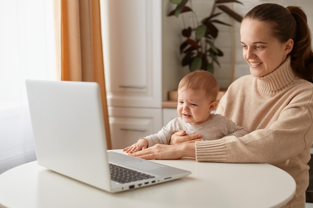포니테일 헤어스타일을 한 행복한 여성의 실내 사진은 캐주얼한 베이지색 스웨터를 입고 어린 아기와 함께 부엌에 있는 테이블에 앉아 긍정적인 표정으로 노트북 디스플레이를 바라보고 있습니다.