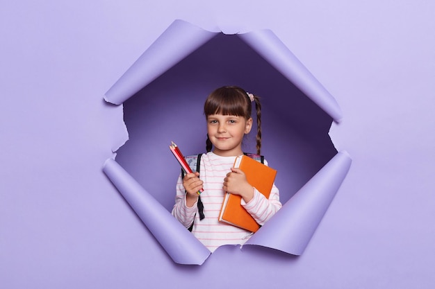 Indoor shot of smiling joyful little girl in a striped shirt with textbooks and pencils in her hands and a backpack schoolgirl with braids posing in paper hole isolated over lilac background