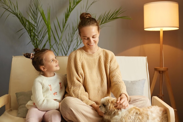 Indoor shot of smiling mother and little daughter playing with dog while sitting on sofa at home child with funny hair buns looking smiling at mommy family enjoying time together