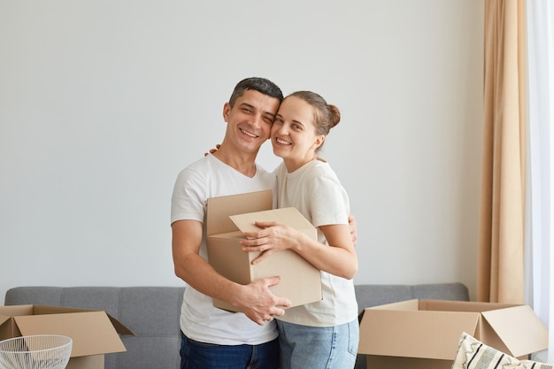 Ripresa in interni di una coppia positiva sorridente in piedi con un pacco di cartone nel nuovo appartamento che si abbraccia e guarda la fotocamera con un viso felice che si esprime soddisfatto di acquistare la propria casa